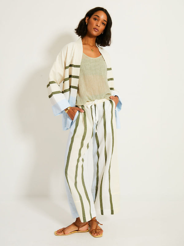Woman Standing Wearing lemlem Desta Pants featuring a bold asymmetrical color-block pattern, where airy light blue effortlessly blends with soft sage and cream, matching jacket and sheer tank in sage color