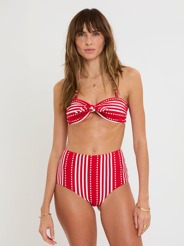 Woman wearing lemlem ELSI High Waist Bottom and AVA Bandeau Top featuring featuring a vanilla ground accented with vibrant red stripes and a dotted pattern. on a plain background