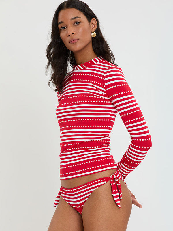 Woman wearing lemlem LUCY Side Tie Bottom and AZEB Rashguard featuring featuring a vanilla ground accented with vibrant red stripes and a dotted pattern. on a plain background