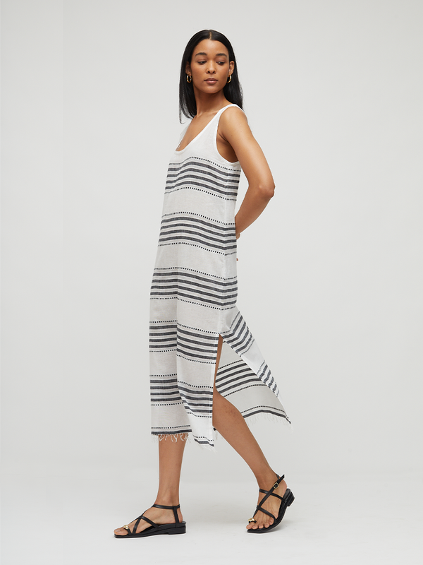 A woman modeling a lemlem LILIAN handwoven sleeveless white knit midi dress with horizontal black stripes and a side slit, paired with black strappy sandals, against a light gray studio background.