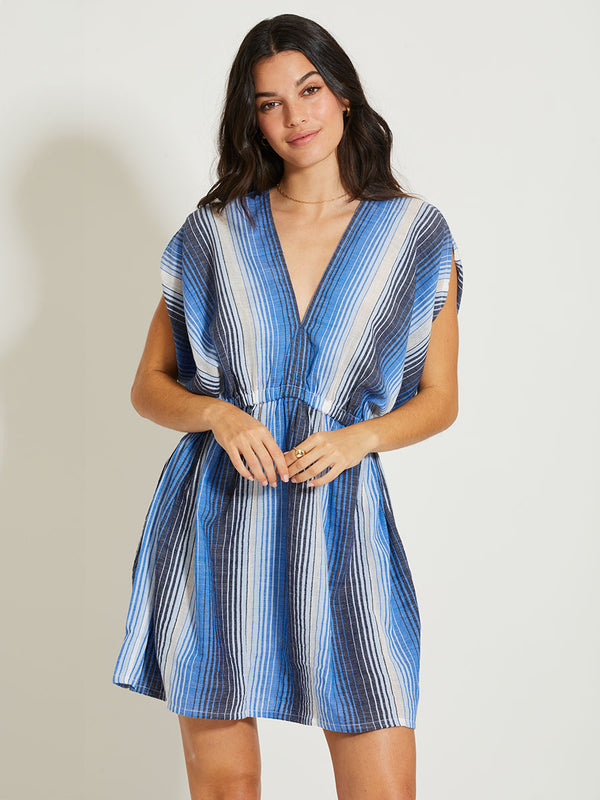 A woman standing against a white background wearing a short, sleeveless, striped blue and white dress with a deep plunge neckline and elastic waist.