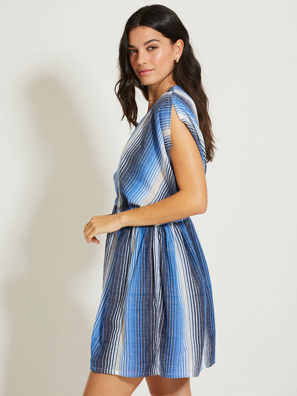 A woman standing against a white background wearing a short, sleeveless, striped blue and white dress with a deep plunge neckline and elastic waist.