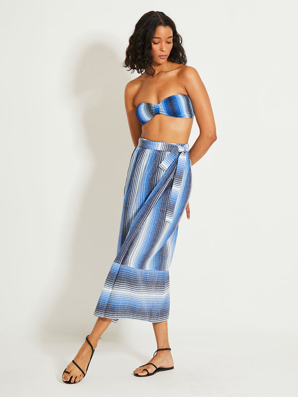 Woman Standing Wearing lemlem SALANA | Wrap Skirt featuring  stripe pattern that blends into a stunning ombré effect, with soft transitions between white, sky blue, mid blue, and deep navy.