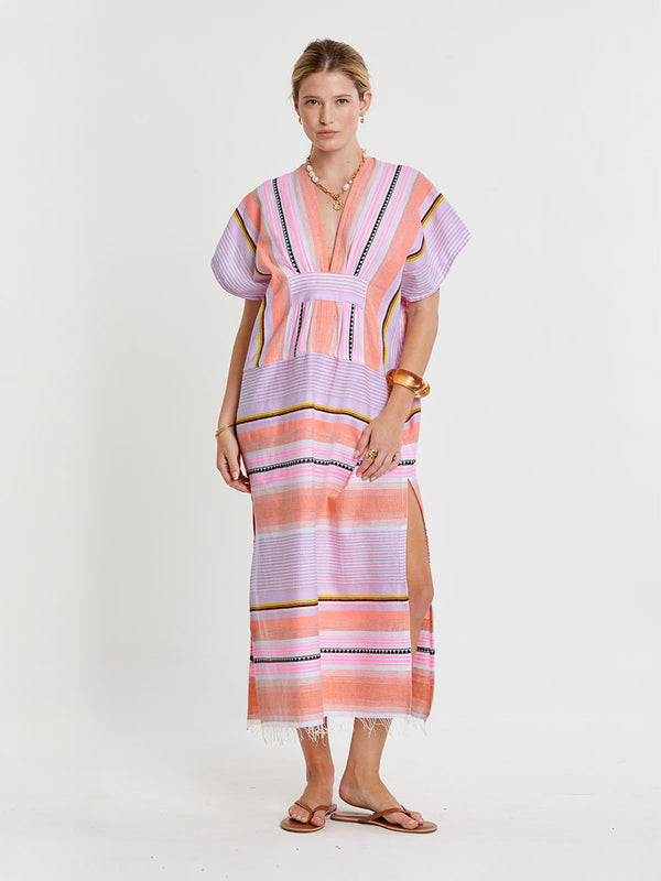 Woman standing wearing lemlem handwoven GASIRA Caftan featuring intricate stripe pattern interlaced with delicate mini Tibeb detailing, expressed in a soft yet vivid palette of cool pink, warm tangerine, rose, orchid, and accents of peach and deep burgundy.