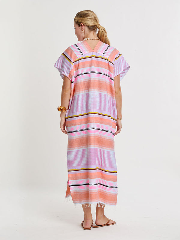 Woman standing wearing lemlem handwoven  GASIRA Caftan  featuring intricate stripe pattern interlaced with delicate mini Tibeb detailing, expressed in a soft yet vivid palette of cool pink, warm tangerine, rose, orchid, and accents of peach and deep burgundy.