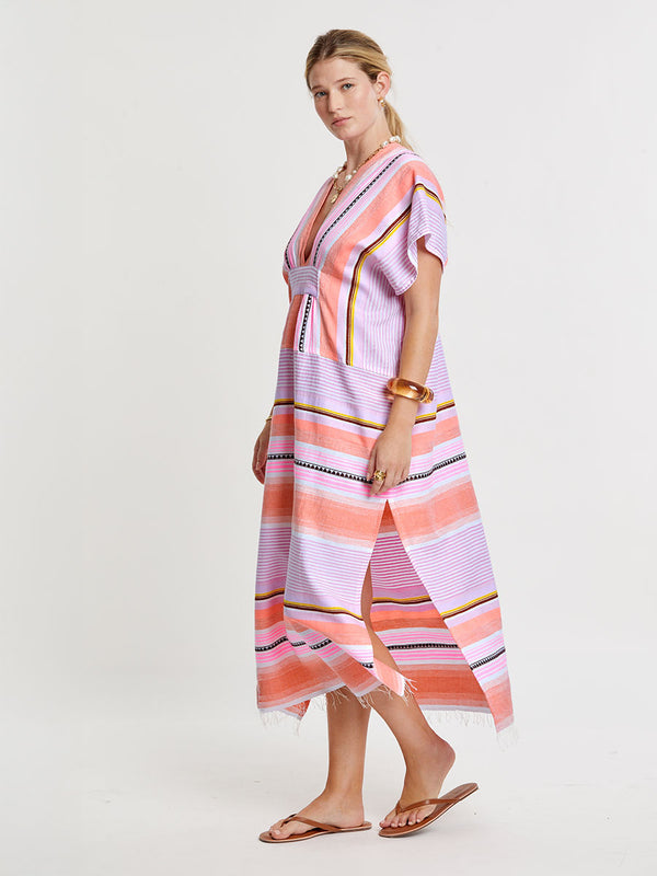 Woman standing wearing lemlem handwoven GASIRA Caftan featuring intricate stripe pattern interlaced with delicate mini Tibeb detailing, expressed in a soft yet vivid palette of cool pink, warm tangerine, rose, orchid, and accents of peach and deep burgundy.