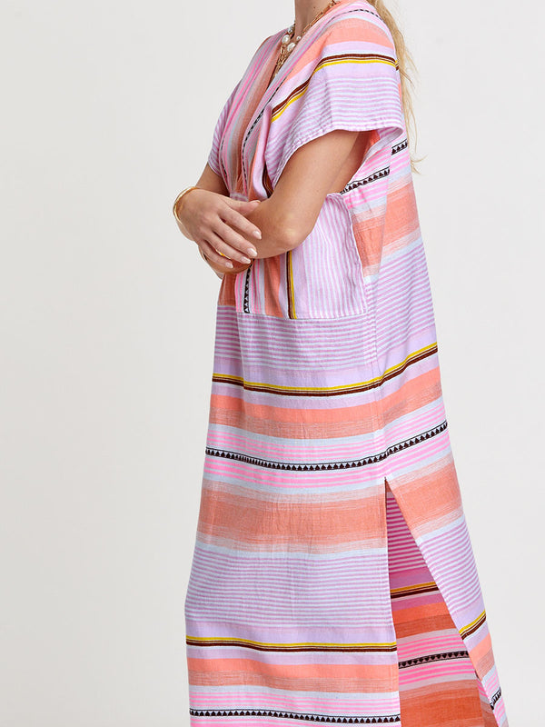 Woman standing wearing lemlem handwoven GASIRA Caftan featuring intricate stripe pattern interlaced with delicate mini Tibeb detailing, expressed in a soft yet vivid palette of cool pink, warm tangerine, rose, orchid, and accents of peach and deep burgundy.