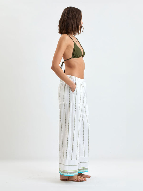 Side Shpt of a  Woman wearing lemlem DESTA Pants  featuring a chic stripe pattern in aqua blue and sage tones, delineated by dark olive dots and accented with a splash of neon yellow on a cream background
