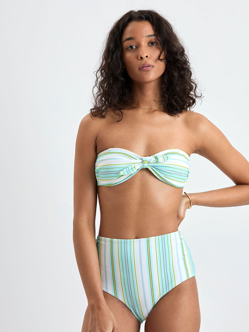 Woman wearing lemlem AVA Bandeau Top and ELSI High Waist Bottom featuring a chic stripe pattern in aqua blue and sage tones, delineated by dark olive dots and accented with a splash of neon yellow on a cream background