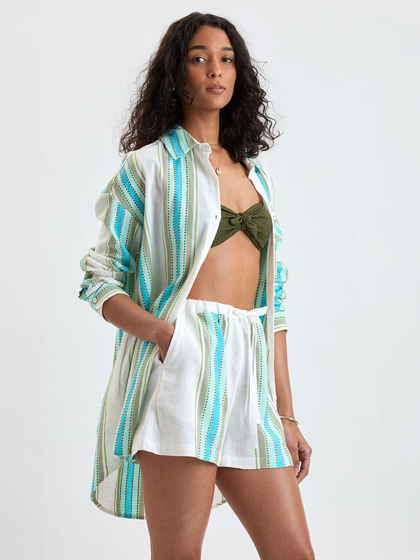 Woman wearing lemlem MARIAM Shirt featuring a chic stripe pattern in aqua blue and sage tones, delineated by dark olive dots and accented with a splash of neon yellow on a cream background