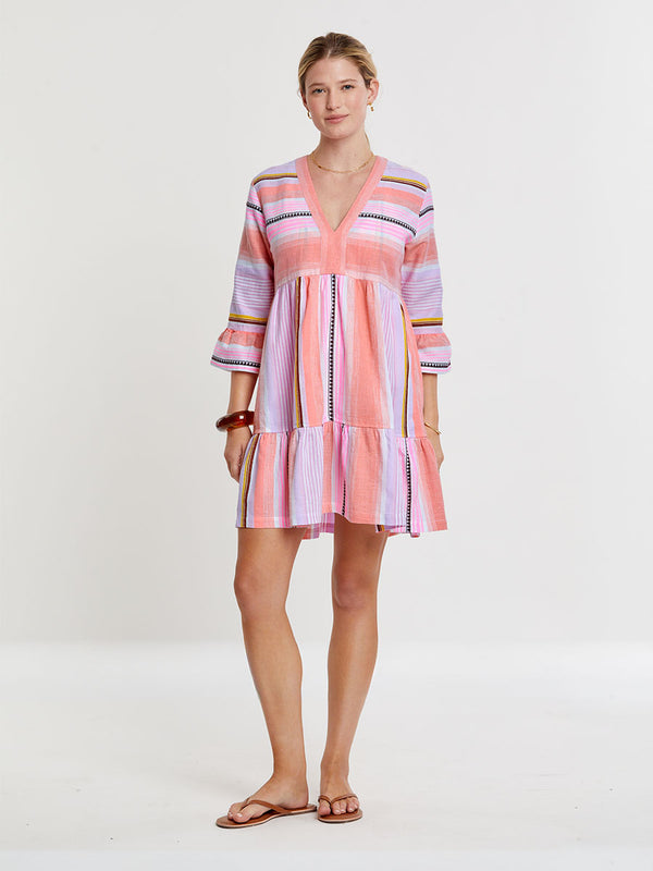 Woman standing wearing lemlem handwoven  HANNA Dress featuring intricate stripe pattern interlaced with delicate mini Tibeb detailing, expressed in a soft yet vivid palette of cool pink, warm tangerine, rose, orchid, and accents of peach and deep burgundy.