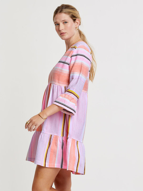 Woman standing wearing lemlem handwoven  HANNA Flutter Dress featuring intricate stripe pattern interlaced with delicate mini Tibeb detailing, expressed in a soft yet vivid palette of cool pink, warm tangerine, rose, orchid, and accents of peach and deep burgundy.