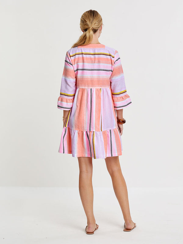 Woman standing wearing lemlem handwoven  HANNA Flutter Dress featuring intricate stripe pattern interlaced with delicate mini Tibeb detailing, expressed in a soft yet vivid palette of cool pink, warm tangerine, rose, orchid, and accents of peach and deep burgundy.