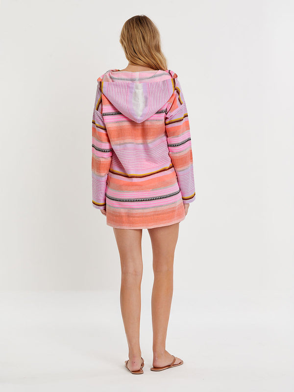 Woman standing wearing lemlem handwoven HENOK Hoodie featuring intricate stripe pattern interlaced with delicate mini Tibeb detailing, expressed in a soft yet vivid palette of cool pink, warm tangerine, rose, orchid, and accents of peach and deep burgundy.