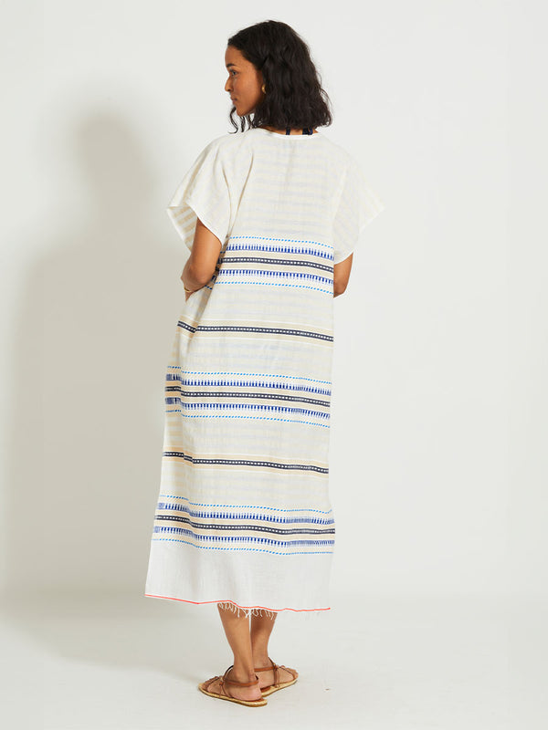 Back of a Woman Standing Wearing lemlem DALILA | V Neck Caftan featuring a soft shadow stripe base with graphic Tibeb-inspired bands in rich blues, black, and vanilla on a creamy white ground