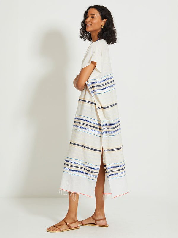 Side of a Woman Standing Wearing lemlem DALILA | V Neck Caftan featuring a soft shadow stripe base with graphic Tibeb-inspired bands in rich blues, black, and vanilla on a creamy white ground