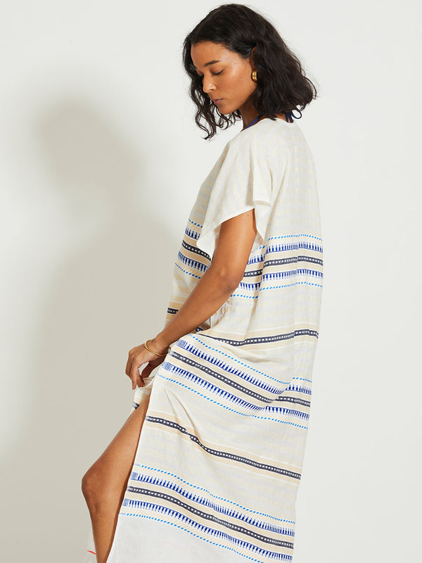 Woman Standing Wearing lemlem DALILA | V Neck Caftan featuring a soft shadow stripe base with graphic Tibeb-inspired bands in rich blues, black, and vanilla on a creamy white ground