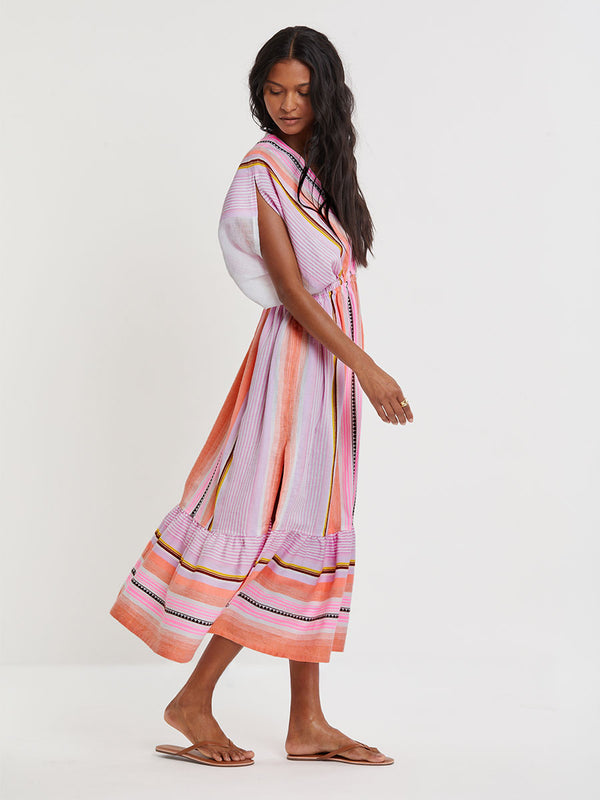Woman standing wearing lemlem handwoven  LEILA Dress featuring intricate stripe pattern interlaced with delicate mini Tibeb detailing, expressed in a soft yet vivid palette of cool pink, warm tangerine, rose, orchid, and accents of peach and deep burgundy.