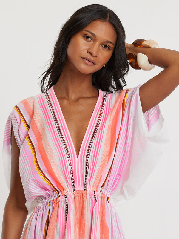 Woman standing wearing lemlem handwoven  LEILA Dress featuring intricate stripe pattern interlaced with delicate mini Tibeb detailing, expressed in a soft yet vivid palette of cool pink, warm tangerine, rose, orchid, and accents of peach and deep burgundy.