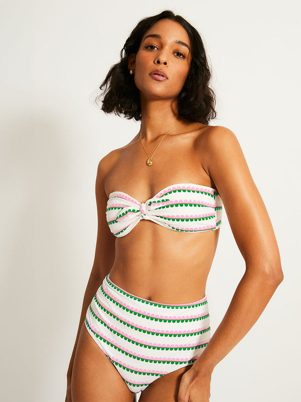 Woman Standing Wearing lemlem AVA Bandeau Top and ELSI High Waist Bottom featuring a playful, preppy scalloped Tibeb weave with vibrant green, soft pink, and fiery red accents.
