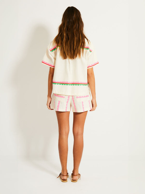 Back of a Woman Standing Wearing lemlem SAFIA Shorts and TUULI Cabana Shirt Top featuring a playful, preppy scalloped Tibeb weave with vibrant green, soft pink, and fiery red accents 
