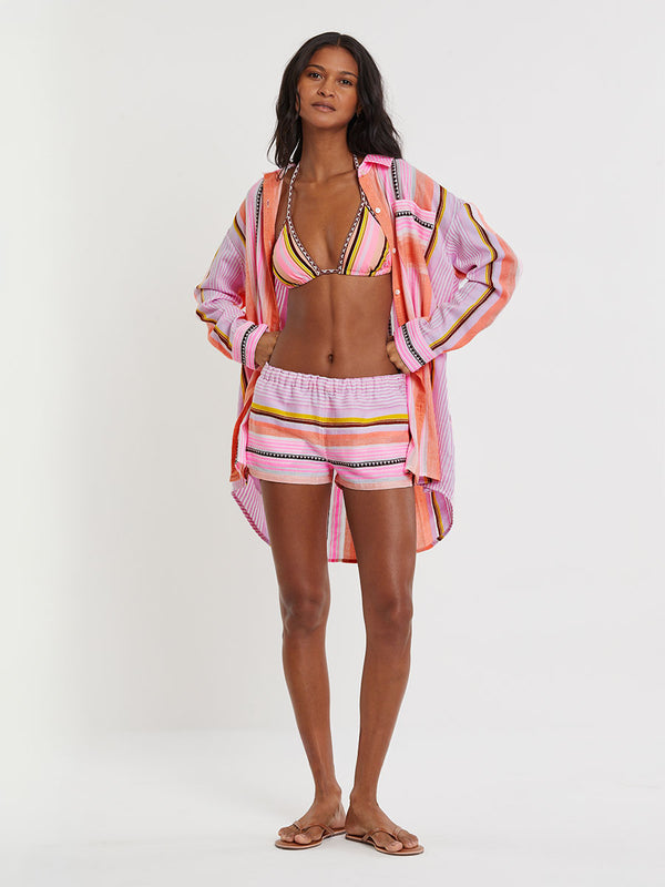 Woman Standing wearing lemlem MALIA Triangle Top and ZENI Shorts featuring intricate stripe pattern interlaced with delicate mini Tibeb detailing, expressed in a soft yet vivid palette of cool pink, warm tangerine, rose, orchid, and accents of peach and deep burgundy.