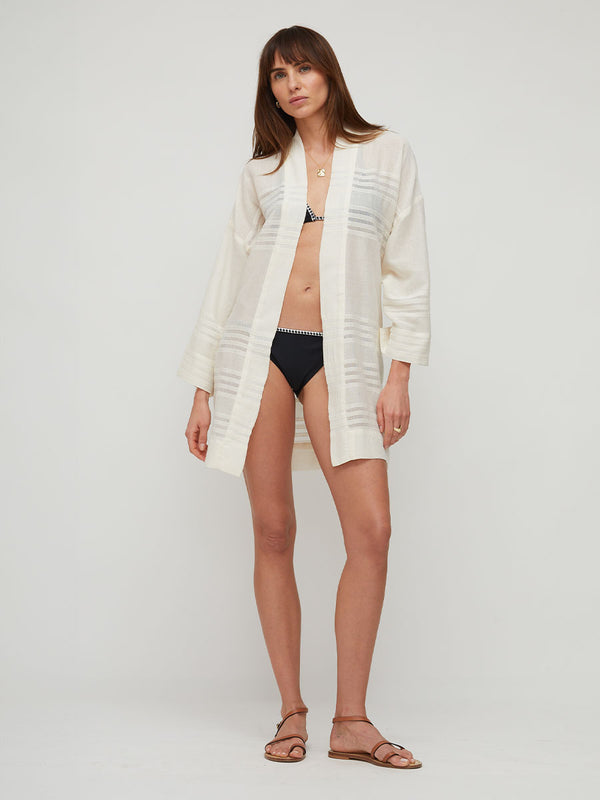 A woman modeling a long-sleeve, open-front cream cotton cover-up with subtle horizontal textured stripes, worn over a black bikini bottom and brown strappy sandals against a light gray studio background.