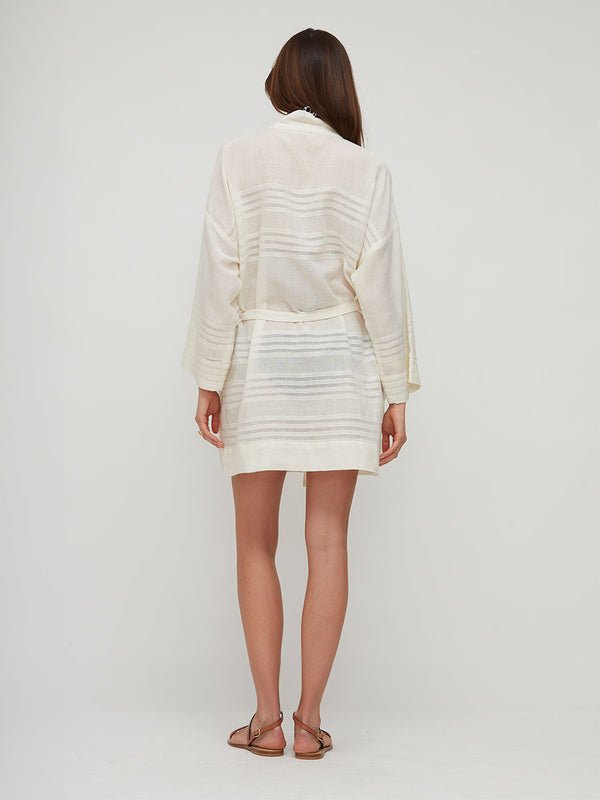 A woman viewed from the back wearing a cream-colored, long-sleeved  lemlem handwoven ALMA Robe with horizontal textured stripes, paired with brown flat sandals, against a plain light gray studio background.