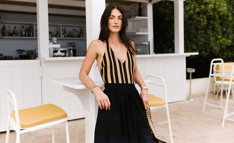 A woman stands outdoors in a white patio setting wearing a sleeveless black and beige vertically striped dress with a deep V-neckline and a black pleated skirt. She is holding a gold-accented clutch.