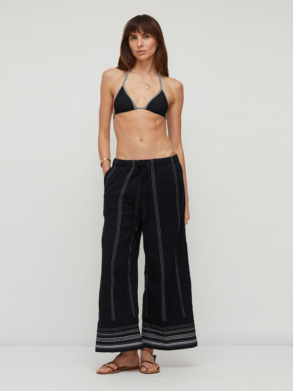 A woman modeling a black two-piece outfit consisting of a halter triangle bikini top with white trim and matching black lemlem handwoven DESTA wide-leg pants with vertical white stripe details and a striped hem, set against a plain light gray studio background.