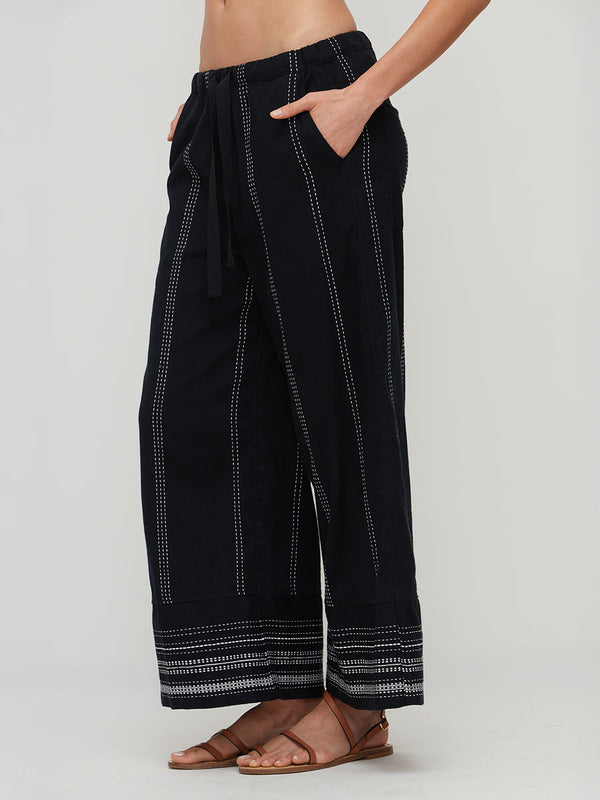 A person wearing lemlem handwoven DESTA  loose black wide-leg pants with vertical dotted stripe details and a striped hem, paired with brown strappy sandals against a light gray studio background.