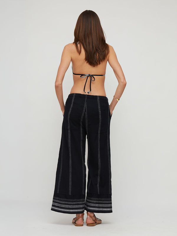 A woman shown from the back wearing wide-leg lemlem handwoven DESTA black pants with vertical and horizontal striped detailing, paired with a black tie-back bikini top, against a plain light gray studio background.