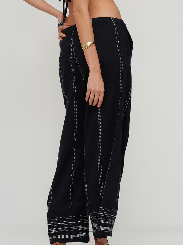 Black wide-leg lemlem handwoven DESTA pants with white embroidered vertical stripes and matching striped hem detailing, shown on a model from the side against a light gray background.