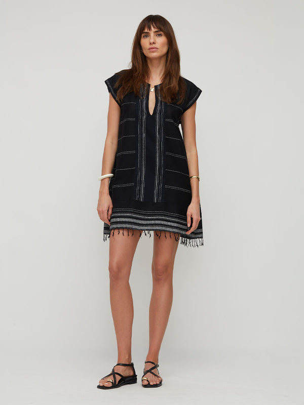A woman modeling a lemlem handwoven ELINA short black dress with white horizontal stripes, a keyhole neckline, and fringe trim, photographed against a plain light gray studio background.