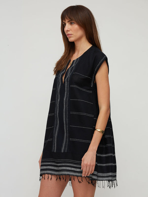 A woman wearing a lemlem handwoven ELINA sleeveless black tunic dress with white dotted horizontal stripes and fringe trim, photographed against a plain light gray studio background.