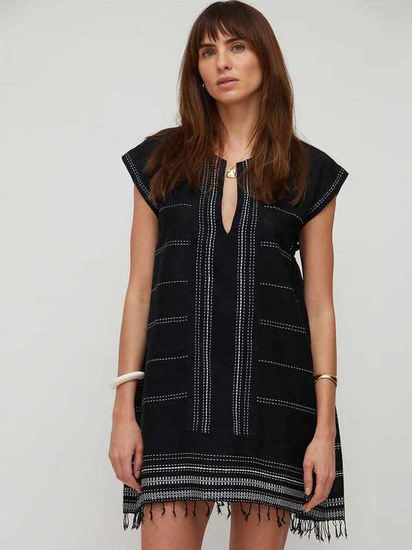 A woman modeling a black lemlem handwoven  sleeveless ELINA tunic dress with white geometric stitched patterns, a split neckline, and fringe trim at the hem, set against a plain light gray studio background.