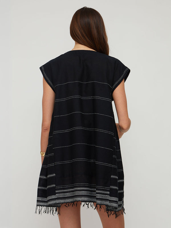 A woman viewed from the back wearing a lemlem handwoven ELINA sleeveless black tunic dress with horizontal white dotted stripe patterns and fringed trim at the hem, against a plain light gray studio background.