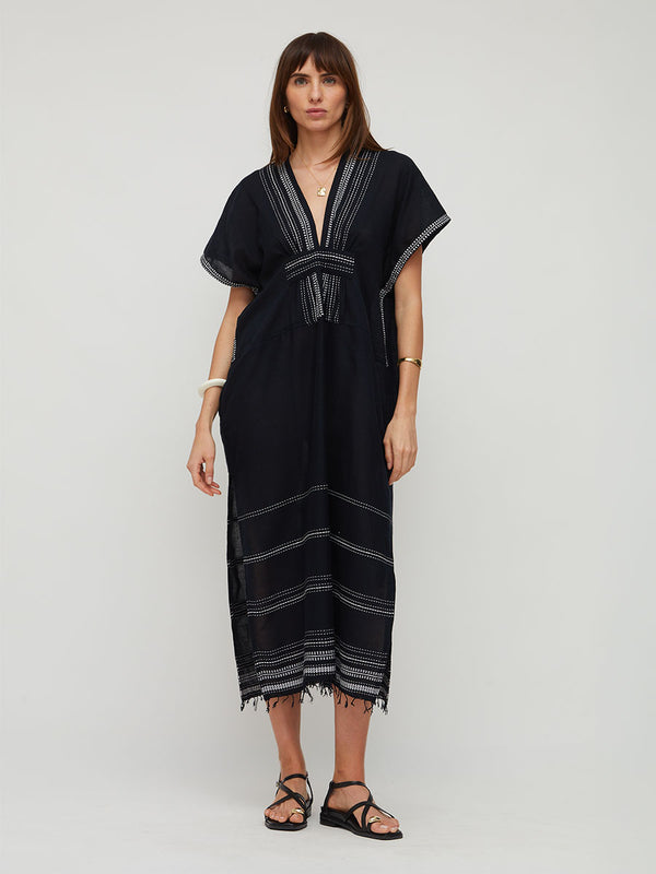 A woman modeling a lemlem handwoven GASIRA black kaftan dress with white striped details and a tie at the waist, paired with black strappy sandals, against a light gray studio background.