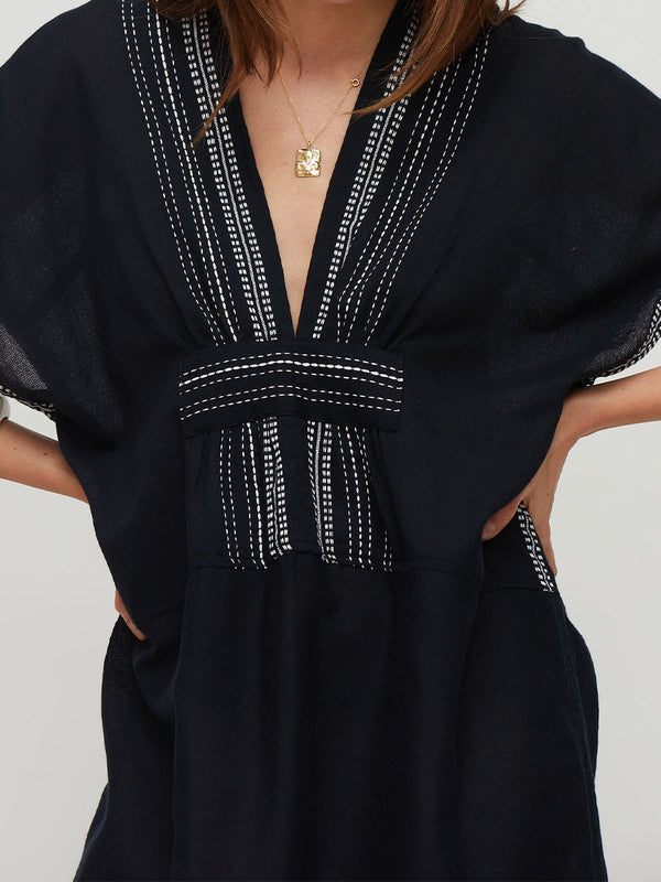 A close-up of a model wearing a black lemlem handwoven GASIRA kaftan dress with white embroidered vertical stripes along the V-neckline and waist tie.