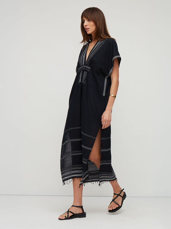 A woman modeling a black midi lemlem handwoven GASIRA kaftan dress with white striped details and a tie waist, paired with black strappy sandals against a light gray studio background.