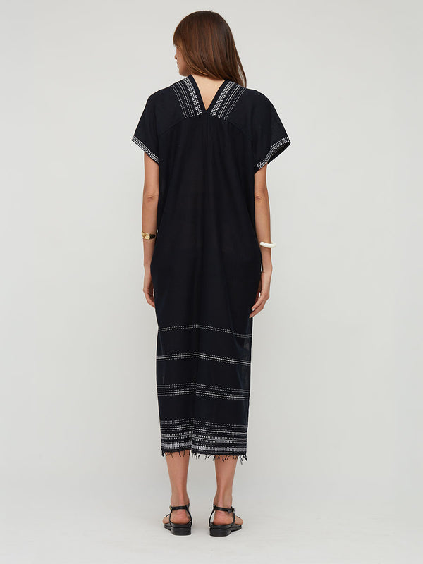 A woman viewed from the back wearing a loose lemlem handwoven DALILA black caftan dress with white striped trim along the neckline, sleeve cuffs, and hem, paired with black sandals against a light gray studio background.