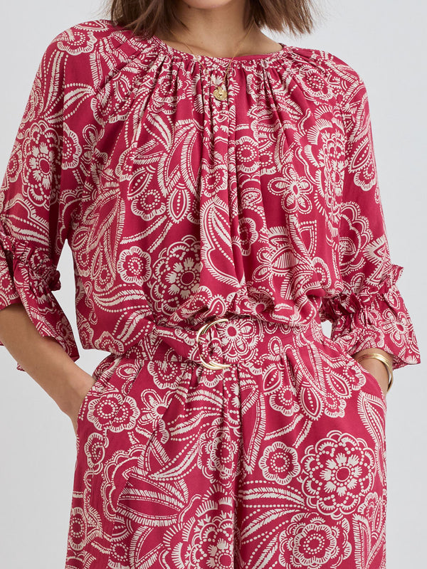Close up on a  Woman Standing Wearing lemlem MONIKA Blouse and DESTA Pants featuring rich ruby-red fabric  adorned with intricate white floral and leaf motifs print