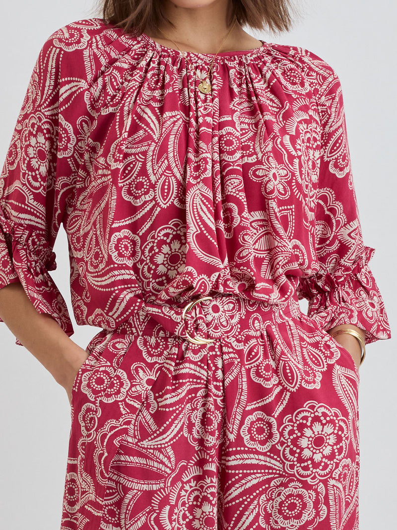 Close up on a  Woman Standing Wearing lemlem MONIKA Blouse and DESTA Pants featuring rich ruby-red fabric  adorned with intricate white floral and leaf motifs print
