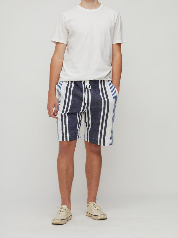 A model wearing lemlem handwoven navy blue and white vertically striped drawstring shorts, a white t-shirt, and beige sneakers, standing against a light gray studio background.