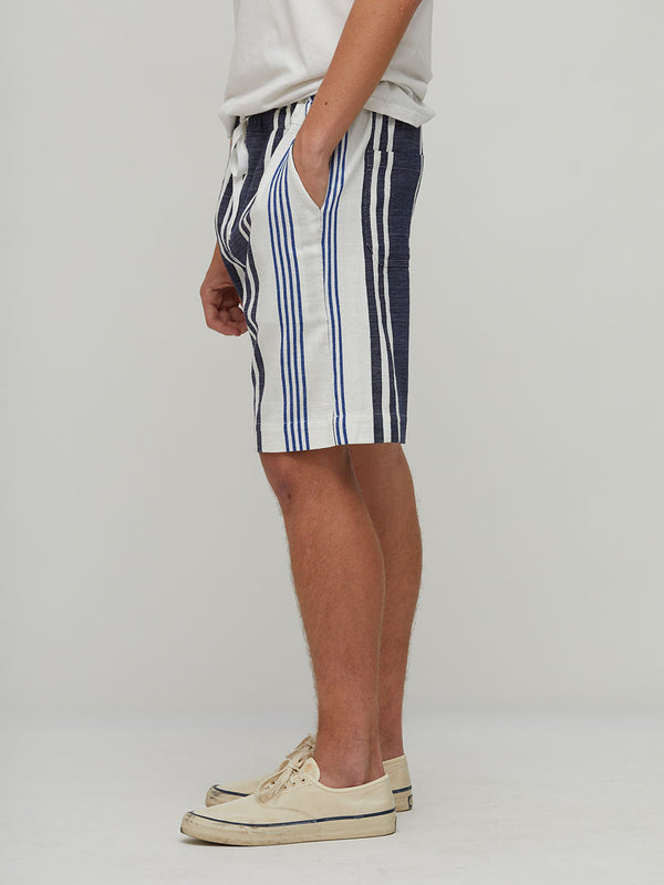 A person shown from the waist down wearing lemlem handwoven striped shorts with vertical navy, blue, and white stripes, paired with a white t-shirt and beige canvas sneakers, against a light gray studio background.