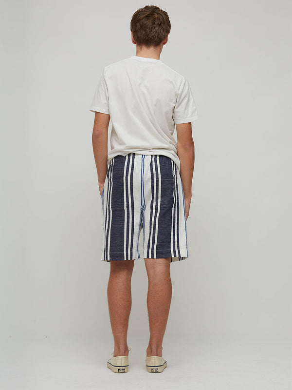 A man viewed from behind wearing a white t-shirt and lemlem handwoven navy blue and white vertically striped shorts, standing against a light gray studio background.