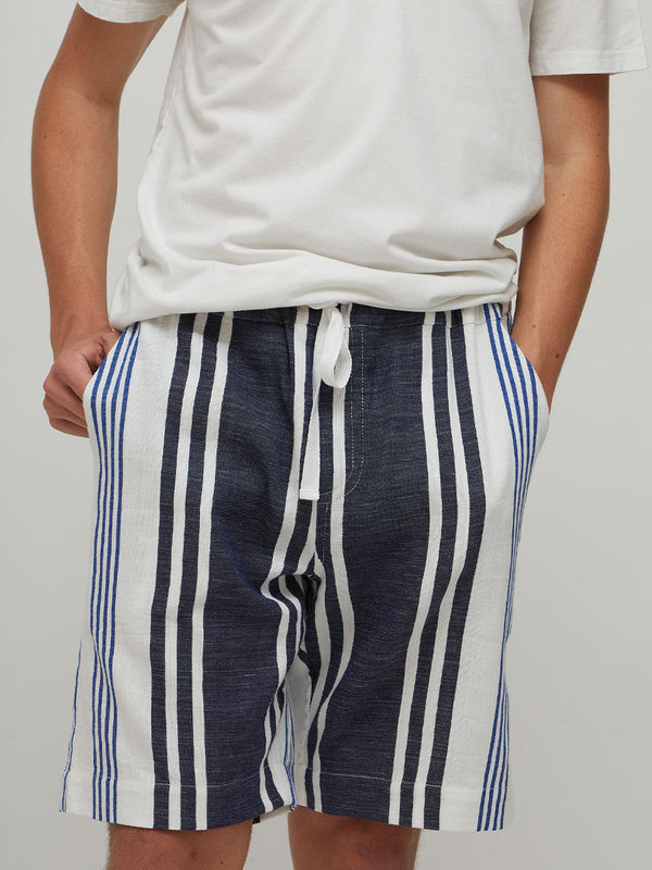 A person wearing lemlem handwoven  navy and white vertically striped drawstring shorts with a white t-shirt, shown from the chest down against a light gray studio background.