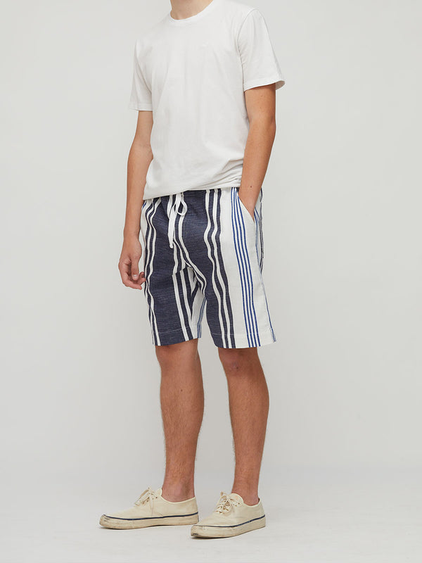 A man wearing lemlem handwoven navy blue and white vertically striped drawstring shorts, a plain white t-shirt, and cream-colored canvas sneakers, standing against a light gray studio background.