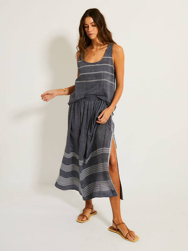 Woman Standing Wearing lemlem Freye Skirt featuring chambray navy color accented with fine, textured white stripes and matching tank top.