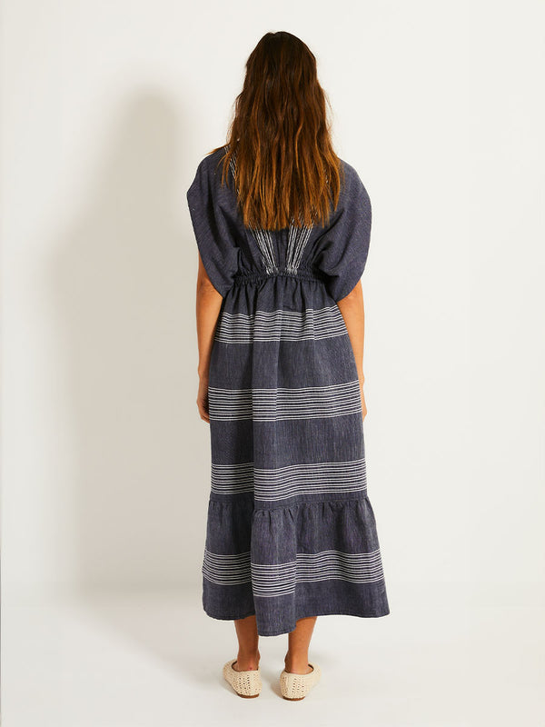 Back of a Woman Standing Wearing lemlem  LEILA Dress  featuring chambray navy color accented with fine, textured white stripes
